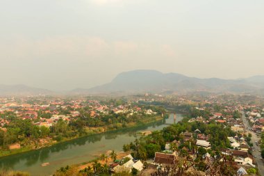 Laos 'un Luang Prabang kentinin geniş açılı perspektifi, Mont Phousi' den Vietnam Han Nehri 'nin şehri sardığı ve arka planda puslu dağların olduğu görülüyor. Görüntü kırmızı çatılı evleri, tropikal yeşillikleri ve nehrin altında sakin bir manzarayı yakalıyor.