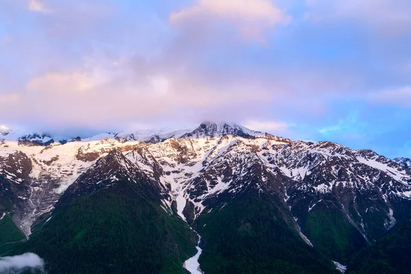 Ilık sabah ışığı, Mont Blanc 'ın engebeli, karla kaplı tepelerini aydınlatır. Altında yeşil ormanlar ve tepesinde pastel bulutlar vardır..