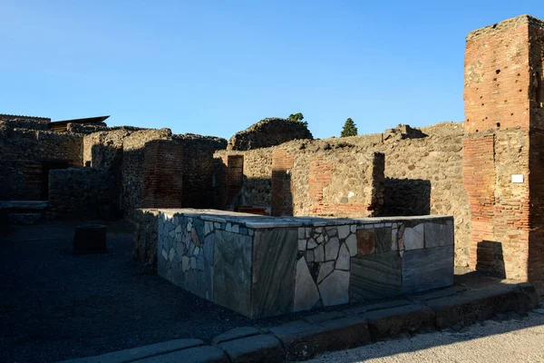 İtalya 'nın Pompeii kentindeki arkeolojik harabelerde yıpranmış tuğla sütunların ve volkanik taş duvarların önünde bir mozaik taş tezgah duruyor. Erken güneş ışığı keskin gölgeler oluşturur, açık bir yüzeyin altındaki dokulu duvarcılık ve geometrik desenleri vurgular.