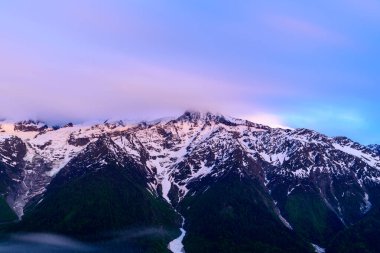 Yumuşak akşam ışığı, Mont Blanc 'ın engebeli, karla kaplı yamaçlarını pembe ve mavi renklerle kaplıyor. İnce bulutlar dramatik zirvelerin üzerinde sürükleniyor..