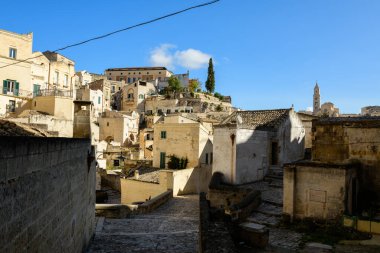 İtalya 'nın Matera kentindeki tarihi kireçtaşı binalar arasında dar kaldırım taşları rüzgarları esiyor. Sıcak güneş ışığı yüzleri aydınlatıyor ve açık mavi gökyüzünün altında uzak bir katedral kulesi var..