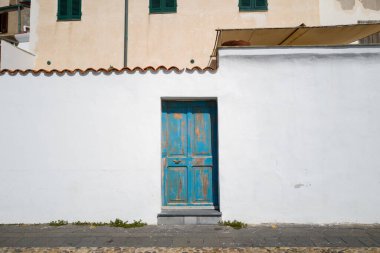 Sardinya, Alghero 'nun tarihi merkezinde parlak beyaz bir sıva duvarına karşı kırsal mavi bir ahşap kapı duruyor. Güneş ışığı, kabuğu soyulmuş boya ve basit dokuları vurguluyor ve bir Akdeniz atmosferi yaratıyor..