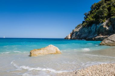 Güneşli bir kaya parçası, bir çakıl taşı plajında, yumuşak turkuaz dalgalarda, engebeli kireçtaşı uçurumlarında ve yemyeşil sardalyalarda, berrak mavi bir gökyüzünün altında yükseliyor..