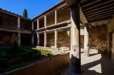 Antik taş sütunlar Pompei, İtalya 'daki Aşıklar Evi' nde geometrik bir bahçe çerçevesi oluşturuyor. Kıtır güneş ışığı, yıpranmış duvarcılık ve manikürlü boxwood çitleri üzerinde güçlü gölgeler oluşturarak Roma mimari tasarımının izlerini ortaya çıkarıyor..