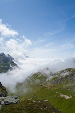 Bereketli yeşil çayırlar, engebeli kaya oluşumları ve berrak mavi gökyüzünün altında vadide sürüklenen alçak bulutlar içeren Beaufortain bölgesinde geniş bir dağ manzarası..