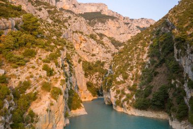 Sıcak güneş ışığı, engebeli kireçtaşından kayalıkları yıkıyor ve Gorges du Verdon 'daki turkuaz bir nehrin üzerindeki yemyeşil bitki örtüsü sakin ve davetkar bir manzara yaratıyor..