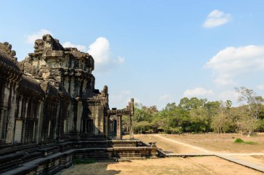 Angkor Vat 'taki yıpranmış bir kumtaşı pavyonu güneş ışığında duruyor. Karmaşık oymaları ve geometrik çizgileri kuru otlar ve açık araziyle çelişiyor. Sembolik Khmer mimarisi dağınık ağaçlar ve soluk mavi bir zemin üzerine kurulmuştur.