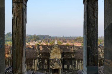 Antik taş sütunlar vasıtasıyla simetrik perspektif Angkor Vat, Kamboçya 'nın büyük giriş ve dış galerilerini ortaya çıkarır. Tarihi tapınak kompleksi kuru topraklar, dağınık palmiye ağaçları ve soluk mavi bir ormanın altındaki uzak ormanlarla çevrilidir.