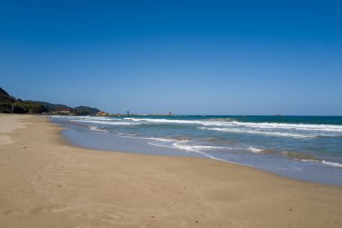 Kumihamacho 'daki Shotenkyo Sahili' nde geniş bir altın kum tabakası dalgalarla karşılaşıyor, canlı mavi gökyüzü ve uzak yeşil tepeler sakin bir sahil manzarası yaratıyor..
