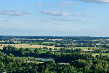 Loire Vadisi 'nin panoramik bir manzarası. Yeşil ve altın tarlalardan oluşan bir mozaik, sık ağaç çizgileri ve yumuşak bulutlarla kaplı bir gökyüzünün altında dolambaçlı bir nehir. Sahne güneşli, huzurlu ve geniş..