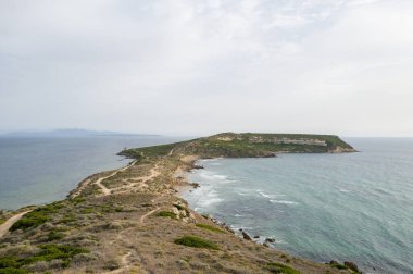 Sardinya 'nın Capo San Marco kentindeki Akdeniz' e uzanan uzun ve dar bir yarımada, kayalık kıyılarla ve yumuşak, bulutlu bir gökyüzünün altındaki seyrek kıyı bitkileriyle sınırlıdır. Dolambaçlı toprak yol ve desenli manzara izolasyon hissi uyandırıyor.