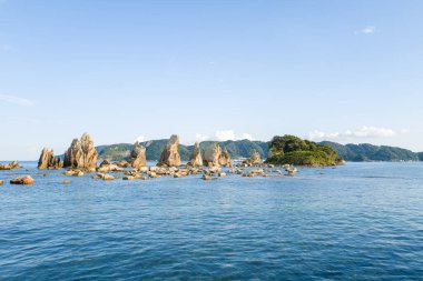 Japonya 'nın Kushimoto kentindeki Hashiguiwa' da yemyeşil bir adanın yanında durgun mavi sulardan yükselen güneşli taş kuleleri ve dağınık kayalar berrak bir gökyüzünün altındaki uzak orman tepeleri ile.