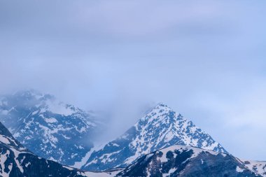 Mont Lachat 'ın işaret ettiği zirve, engebeli yamaçlarda eriyen kar çizgileriyle sürüklenen sisin içinden çıkıyor. Yumuşak mavi ışık ve bulutlu gökyüzü sakin, ruhani bir alp havası yaratır..