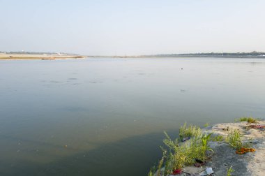 Prayagraj, Hindistan 'daki Ganj ve Yamuna nehirlerinin kesiştiği yerde sessiz bir sahne. Yumuşak su, dağınık yeşillik ve açık gün ışığında sunulan adaklarla dolu kumlu bir kıyıya rastlar..