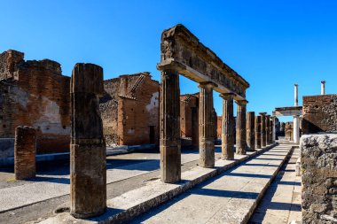 Bir sıra yıpranmış taş sütun ve tuğla duvarlar Pompeii, İtalya 'da bir idari binanın kalıntılarını oluşturuyor. Parlak güneş ışığı, dokulu duvarlar boyunca keskin gölgeler bırakır ve parlak mavi gökyüzünün altında açık bir yürüyüş yolu açar..