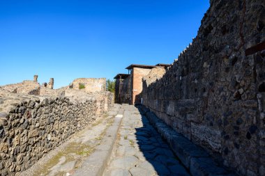 Pompeii 'nin arkeolojik bölgesinde, canlı mavi gökyüzünün altında yıpranmış taş ve tuğla duvarlarla kaplı dar bir cadde. Görüntü, bu Roma şehrinin dokulu yapısını, keskin gölgelerini ve tarihi şehir planlarını vurguluyor..