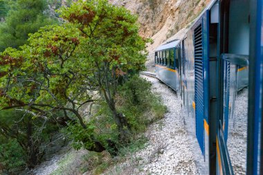 Yunanistan 'ın Kalavrita kenti yakınlarındaki Vouraikos Vadisi' nde kayalık yamaçlar ve yoğun yeşil ağaçlarla çevrili engebeli bir dağ manzarasında mavi dar ölçülü bir tren esiyor. Sahne, sarp kayalıklarla gür yapraklar arasındaki zıtlığı yakalar.