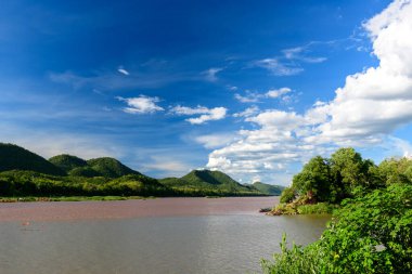 Çamurlu Mekong nehrinin geniş manzarası yemyeşil tepeleri ve Luang Prabang, Laos yakınlarındaki yoğun nehir kenarı bitkilerini geçiyor. Dağınık bulutlu parlak mavi gökyüzü seyahat ve doğa kavramları için ideal canlı, doğal bir manzara yaratır..