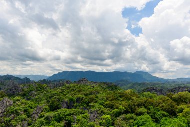Dalgalanan bulutlarla dolu bir gökyüzünün altındaki yoğun yeşil ormandan sivri kireçtaşı oluşumları yükseliyor. Sahne, yağmur mevsiminde Laos kırsalının tipik engebeli arazisini ve yemyeşil bitki örtüsünü yakalıyor..