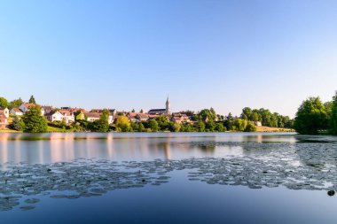 Yüzen zambaklar, Dampierre en Burly 'nin çatılarını ve kilise kulesini yansıtan durgun bir göletin yüzeyini kaplıyor. Sıcak güneş ışığı ve berrak mavi gökyüzü huzurlu kırsal manzarayı güçlendirir..