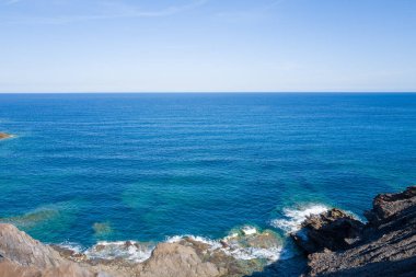 Açık mavi Akdeniz suları Arjantin yakınlarındaki uzak sahil şeridi boyunca kayalıklarla kesişiyor. Güneş ışığı, bulutsuz bir gökyüzünün altındaki canlı deniz tonlarını ve dokulu taşları vurgular..