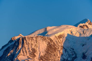 Sabahın erken saatlerinde kar kaplı zirvede yıkanır ve Mont Blanc 'ın engebeli kaya yüzleri ılık turuncu tonlarda, çarpıcı bir alp kontrastı için koyu mavi gökyüzüne vurur..