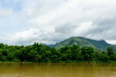 Çamurlu Nam Ou Nehri boyunca uzanan yeşil tepeler, katmanlı bulutlarla dolu bir gökyüzünün altında arka planda yükseliyor. Sahne, Kuzey Laos 'un yemyeşil bitki örtüsünü ve sakin atmosferini yakalıyor..