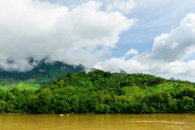 Kuzey Laos 'taki Muang Ngoi yakınlarındaki çamurlu Nam Ou nehri boyunca yemyeşil tepeler ve yoğun tropikal ormanlar üzerinde yoğun bulutlar gezinir. Hareketli manzara kıvrımlı yamaçlar ve dramatik bir gökyüzü içeriyor, sakin bir nehir kenarı manzarası yaratıyor..