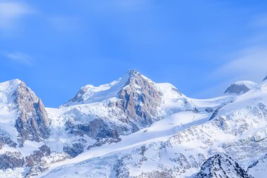 Mont Maudit 'in engebeli, karla kaplı zirvesi, parlak mavi gökyüzüne karşı keskin bir şekilde yükselir, buzlu yamaçlar ve çıplak kaya yüzeyleri çarpıcı alp zıtlıkları yaratır..