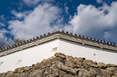 Engebeli bir taş temelin üzerinde yükselen geleneksel fayanslarla el değmemiş beyaz alçıdan kale duvarının açısal görüntüsü, canlı mavi gökyüzünde dağınık bulutlardan oluşan bir zemin üzerine kurulmuş..