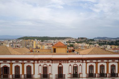 Kemerli pencereleri ve terracotta fayanslarıyla dekoratif tarihi bina, yumuşak bulutlu bir gökyüzünün altında uzak yeşil tepeleri olan Cagliari kentsel manzarasına bakıyor..