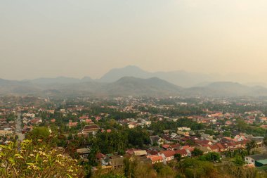 Luang Prabang, Laos 'un yüksek manzarası, kırmızı kiremitli çatıları ve yemyeşil verimli tropikal arazisi uzak sisli tepelere doğru uzanıyor. Yumuşak, altın bir gün ışığı şehrin ve dağların üzerinde sakin bir atmosfer yaratır..