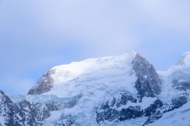Yumuşak, yayılmış güneş ışığı, kar kaplı zirveyi ve Mont Blanc du Tacul 'un engebeli buz oluşumlarını, sakin mavi gökyüzü ve ince bir sis ile nazikçe aydınlatır..