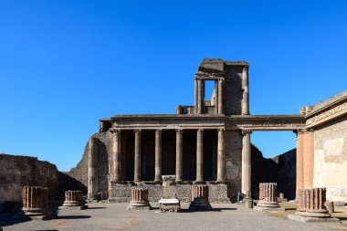 İyi korunmuş taş sütunlar ve antik duvarcılık, İtalya 'nın Pompeii arkeoloji sahasındaki bazilikanın kalıntılarını oluşturur. Parlak güneş ışığı geometrik çizgileri ve dokulu yüzeyleri canlı, bulutsuz bir gökyüzüne karşı güçlendirir..