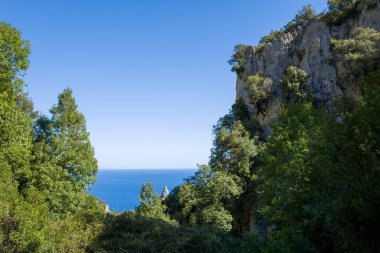 Dik kireçtaşı kayalıkları Akdeniz 'in yoğun ormanlarının üzerinde yükseliyor. Arkasında, Sardunya, Baunei yakınlarındaki derin mavi denizi bir anlığına görüyorlar. Parlak güneş ışığı berrak gölgeler bırakır ve berrak gökyüzünün altındaki yeşil yeşil yaprakları vurgular..