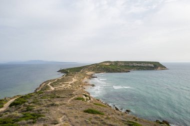 Capo San Marcos 'un açık turkuaz deniz ve yumuşak, bulutlu bir gökyüzü altında engebeli kıyı bitkileriyle çevrili rüzgarlı yarımadasının geniş manzarası. Doğal toprak kıyı şeridi boyunca daha dolambaçlı yol alır, izolasyon ve vahşi güzellik hissi yaratır..