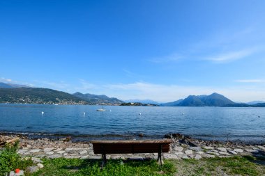 Orman tepeleri ve dağlık tepeler tarafından çerçevelenmiş Isola dei Pescatori silueti ve uzak yelkenlisi ile, Maggiore Gölü 'nün sakin mavi sularına bakan ahşap bir bank. Göl kıyısı sahnesi kuzey İtalya 'da geniş ve temiz bir gökyüzünün altında çekilmiştir..