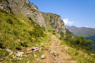 Pireneler 'deki yüksek uçurumların altındaki çimenli yamaçlardan ve kayalık çıkıntılardan geçen engebeli bir yürüyüş yolu açık mavi gökyüzünün altında sıcak güneş ışığı altında yıkanır..