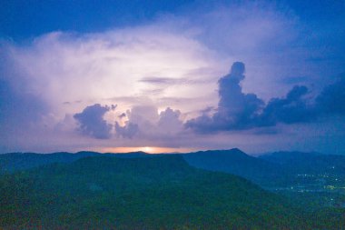 Karanlık çöktüğünde ormanlık bir dağ sırasının üzerindeki uzak yıldırımlar tarafından aydınlatılan karanlık fırtına bulutlarının geniş bir görüntüsü. Sahne, katmanlı siluetleri, parlayan gökyüzünü ve yaklaşan fırtınanın atmosferik gerilimini yakalar..