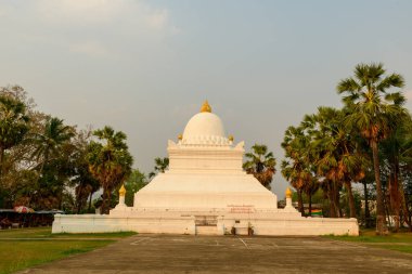 Louang Prabang, Laos 'ta palmiye ağaçları arasında altın kaplamalı beyaz bir stupa duruyor. Sıcak gün ışığı anıtları aydınlatıyor. Temiz çizgiler ve tapınağın çevresindeki tropikal yeşillikler..