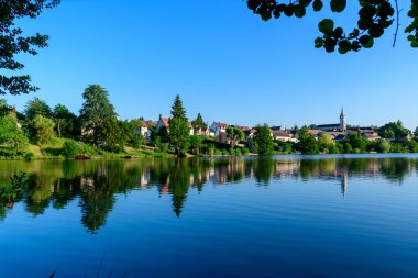 Gölün berrak mavi suyu, Dampierre en Burly 'nin canlı bir yaz gökyüzü altında bulunan çatılarını, kilise kulesini ve bereketli ağaçlarını yansıtır. Asılı dallar sakin kırsal alanı çerçeveletip sakinlik duygusunu artırıyor..