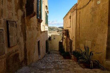 Matera, İtalya 'da sakin, güneşli bir kaldırım taşı sokağı, berrak mavi gökyüzünün altındaki gözlere doğru uzanan yıpranmış taş cepheler ve yeşil saksı bitkileriyle doludur..