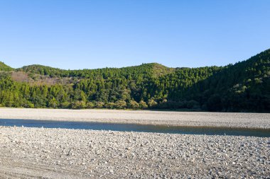 Japonya 'nın Wakayama bölgesindeki Hongu bölgesinde açık mavi bir gökyüzünün altında yoğun ormanlık yeşil tepelerin altından geçen sığ bir deresi olan geniş taşlı nehir kıyısı. Manzara açık ve güneşli, sakin ve doğal bir basitlik hissi uyandırıyor..