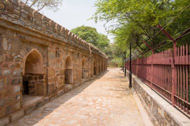 Delhi 'deki Mehrauli Arkeoloji Parkı' nda kemerli nikçikli güneşli taş atari salonu kaldırım hattında. Tarihi duvar, demir çitlerle ve berrak gökyüzünün altındaki yoğun yeşil ağaçlarla çelişiyor..