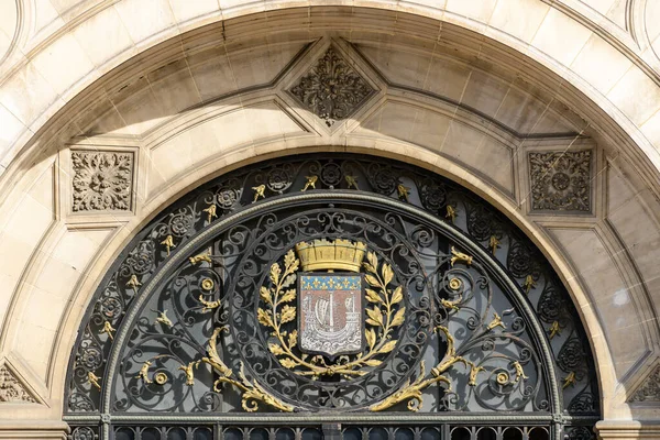 Altın aksanıyla işlenmiş zarif demircilik Paris 'teki Hotel de Ville' deki büyük taştan kemerin altındaki detaylı bir sivil armayı çerçeveliyor. Gün ışığıyla aydınlatılıyor ve karmaşık işçilik sergiliyor..