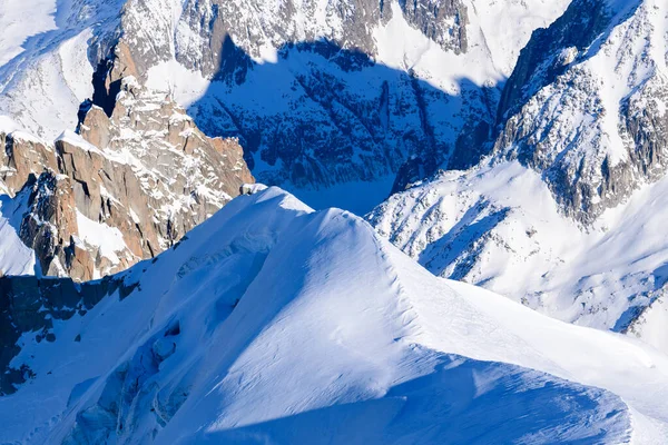 Parlak güneş ışığı, engebeli granit tepeler ve koyu mavi alp gölgelerine karşı koyan Mont Blanc kitlesinde keskin bir kar tepesini aydınlatır..