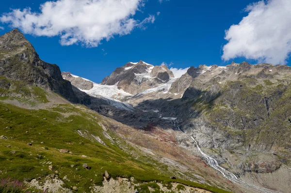 Geniş bir alp manzarası. Buzul de la Lee Blanche, engebeli Aiguilles de Tre la Tete, yemyeşil yamaçlar, ve dağınık mavi bulutların altında çağlayan bir şelale..