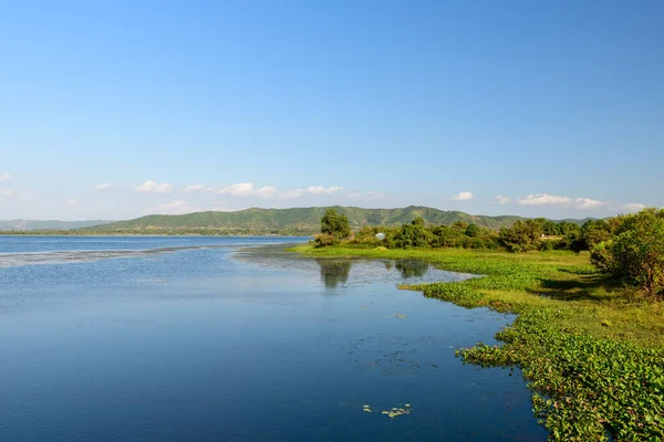 Brateak Krola Gölü 'nün durgun suları açık mavi gökyüzünü ve yemyeşil bitki örtüsünü yansıtır ve uzak tepeler doğal bir zemin oluşturur. Su bitkileri ve dağınık ağaçlar gün ışığında huzurlu kıyı şeridi boyunca uzanır..