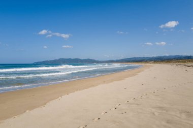 Kumihamacho 'daki Shotenkyo Sahili boyunca uzanan el değmemiş bir kum tabakası, açık mavi gökyüzünün altında hafif dalgalar ve uzak yeşil dağlarla çevrelenmiş..