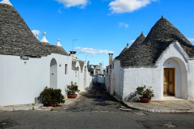 Geleneksel beyaz boyalı trullo evleri konik taştan çatıları olan Alberobello, Apulia, İtalya 'da dar bir şerit çizerler. Saksı bitkileri ve dokulu kireçtaşı duvarlar dağınık bulutlu parlak mavi gökyüzünün altında kendine özgü bir Akdeniz köyü manzarası yaratır.
